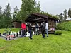 Personer samlet foran en trehytte på en gressplen med utstyr i forgrunnen.