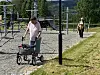 Person med rullator går på en grussti i en park, med lekeapparater og en annen person i bakgrunnen.