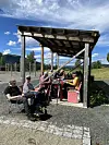 Flere personer sitter under et tak av tre og metall ved et utendørs bord i solskinn.