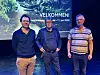 Tre menn står på en scene foran et blått velkomstbanner med tekst og naturmotiv.