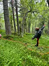 Et barn i turklær balanserer på en line i skogen omgitt av trær og tett grønt vegetasjon.