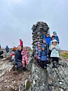 Turkledde og smilende barn og voksne står og sitter på steiner ved en høy varde på en fjelltopp.
