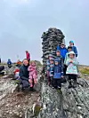 Turkledde og smilende barn og voksne står og sitter på steiner ved en høy varde på en fjelltopp.