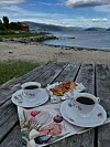 To kopper kaffe og bakverk på et trebord ved en strand med fjord og fjell i bakgrunnen.
