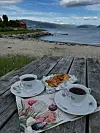 To kopper kaffe og bakverk på et trebord ved en strand med fjord og fjell i bakgrunnen.