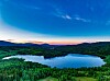 Oversikt over en innsjø omgitt av grønne skoger og åser ved solnedgang.