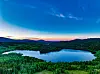 Oversikt over en innsjø omgitt av grønne skoger og åser ved solnedgang.