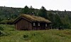 Tømmerhytte med gresskledd tak i en grønn eng ved skogkledd ås.