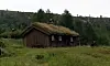 Tømmerhytte med gresskledd tak i en grønn eng ved skogkledd ås.