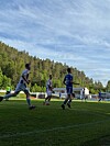 Fotballspillere løper på en gressbane med tribune, skog og blå himmel i bakgrunnen.