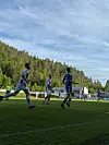 Fotballspillere løper på en gressbane med tribune, skog og blå himmel i bakgrunnen.