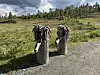 To utskårne treskulpturer ved en grusvei i fjellandskap med gress, trær og overskyet himmel.