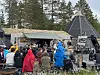 Mennesker (mange av de med jakke med hetten på hodet) samlet ved en lavvo i skogkledd område.