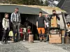 Fire personer står fremst med rekvisitter utendørs. Musikkinstrumenter/utstyr bak.
