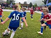 Barn spiller fotball på en gressbane med publikum og telt i bakgrunnen.