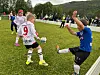 Unge fotballspillere i hvite og blå drakter spiller kamp på en gressbane med publikum i bakgrunnen.