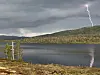 Lyn slår ned over et fjellvann med skogkledd fjell i bakgrunnen under mørke skyer.