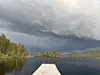 Brygge mot rolig fjellvann med skogkledd strand og mørke skyer på himmelen.
