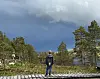 En gutt står på en treplatting i skogen med trær og fjellvann i bakgrunnen. Skyer.