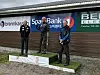 Tre personer står på en seierspall foran sponsorlogoer ved en utendørs arena.