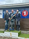 Tre menn på en seierspall ved en skytebane, én holder et gevær.