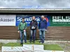 Tre personer står på en seierspall foran en vegg med sponsorlogoer.