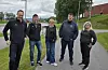 Fem personer står samlet på en grusvei foran gress, trær og en rød bygning.