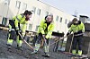 Tre arbeidere i refleksklær arbeider med asfalt ved en bygning.