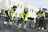 Tre arbeidere i refleksklær arbeider med asfalt ved en bygning.