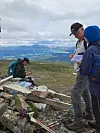 Turgåere står og sitter ved en varde på et fjellplatå med utsikt mot dal og fjell i bakgrunnen.