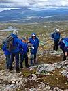 En gruppe turgåere i blå jakker står på et steinete høyfjellsområde med fjell og vann i bakgrunnen.