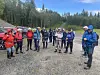 En gruppe personer står samlet med tursekker ved skogkanten under en blå himmel.
