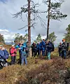 Flere personer står samlet i et skogholt med høye trær og lyng under en grå himmel.