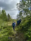 Turgåere med sekker går på en smal skogssti omgitt av trær og busker.