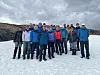 En gruppe mennesker står tett samlet på snø med fjell og skyet himmel i bakgrunnen.
