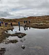Personer går over steinete terreng ved et lite fjellvann under grå skyer.