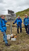 Tre vandrere ved et treskilt i et gresskledd fjellandskap under grå himmel.