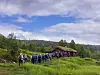 En stor gruppe mennesker går gjennom grønn eng mot en liten hytte i et åpent, kupert landskap.