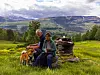 To personer sitter ved en steinrøys med en hund i et grønt fjellandskap.