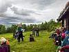 Personer samlet i en gresslette med fjell og skog i bakgrunnen under en skyet himmel.