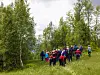 En gruppe mennesker går og samles på en eng i skogkledd terreng med trær og gress.