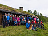 En gruppe personer står og sitter foran en torvtakshytte i fjellet, med to hunder i forgrunnen.