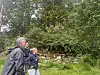 Tre personer går ved en steinmur med tett grønn vegetasjon i en hage.