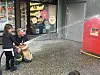 To barn spruter vann fra en slange mot en rød postkasse ved en butikkfasade.