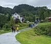 Turgåere går langs en vei gjennom grønt landskap med hus, trær og skogkledde åser i bakgrunnen.