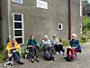 Fem eldre personer i rullestoler sitter ute foran en bygning med skiltet Kallastu.