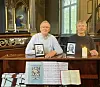 To menn sitter ved et kirkebenk i en kirke med bilder, lys og en åpen bok foran dem.
