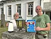 To personer står foran en kirke og holder en plakat ved siden av en stor lykt og en hvit beholder.