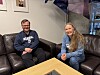 To personer sitter i hver sin sofa i et lyst rom med kaffebord og innrammede bilder på veggen.