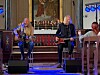 To musikere opptrer med gitar, bass og mikrofoner foran alteret i et kirkerom med blå scenelys.